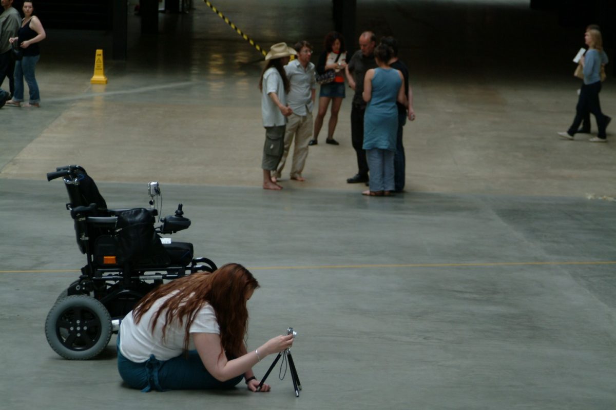 Image ID: A feminine figure sits on the ground in the bottom left of the photograph. They have long reddish/brown hair, wearing a white t-shirt and teal coloured trousers. They are looking through a camera on a tripod, beside them is a wheelchair. Six people are standing in a semi circle in the background and there are other people to the left and right of the photo.