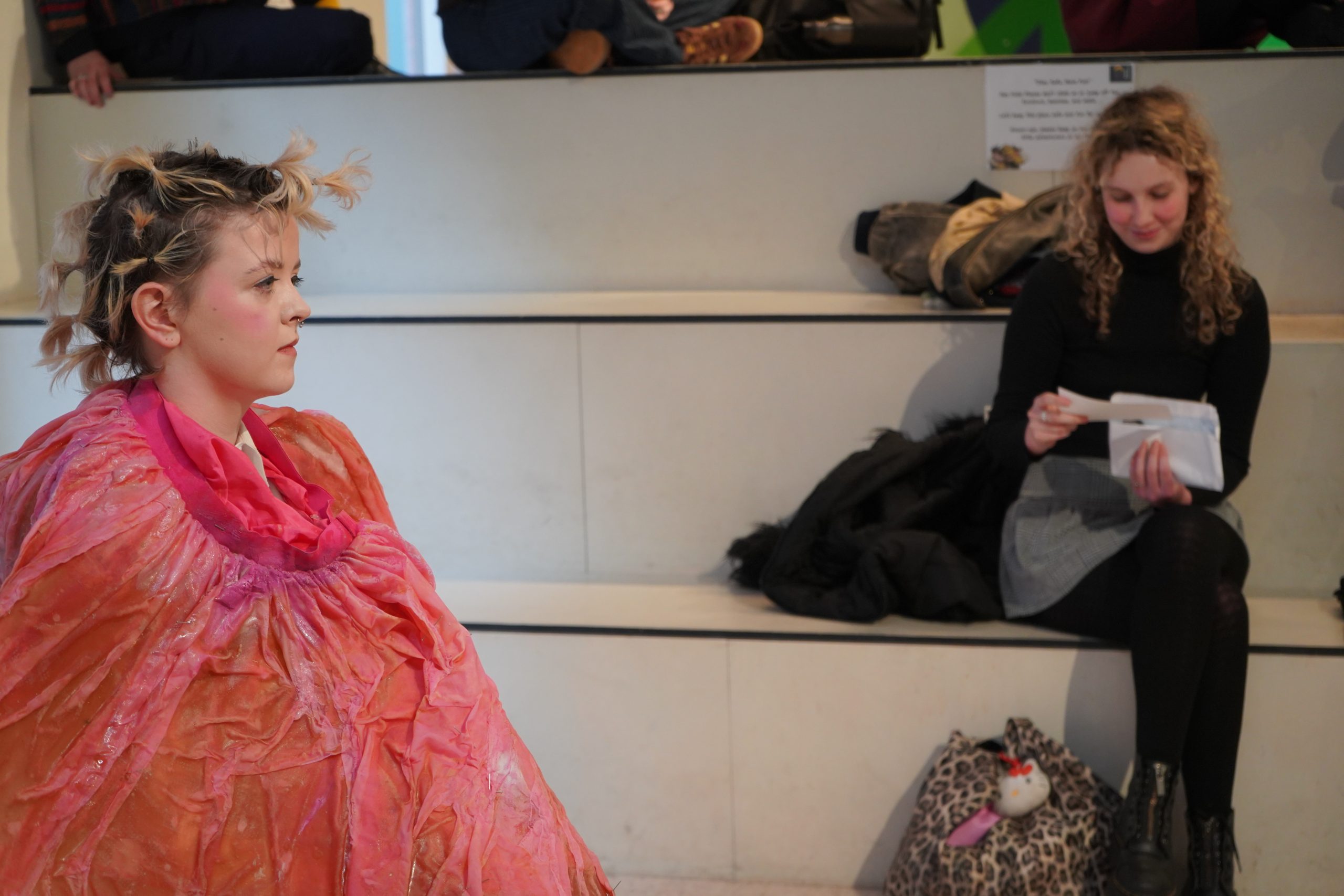 Photograph of an artist sitting in profile. They are wearing a red-pink smock which covers their whole body, and their short hair is in a number of very small bunches over their head. Behind them another person is sitting on steps reading from a page.
